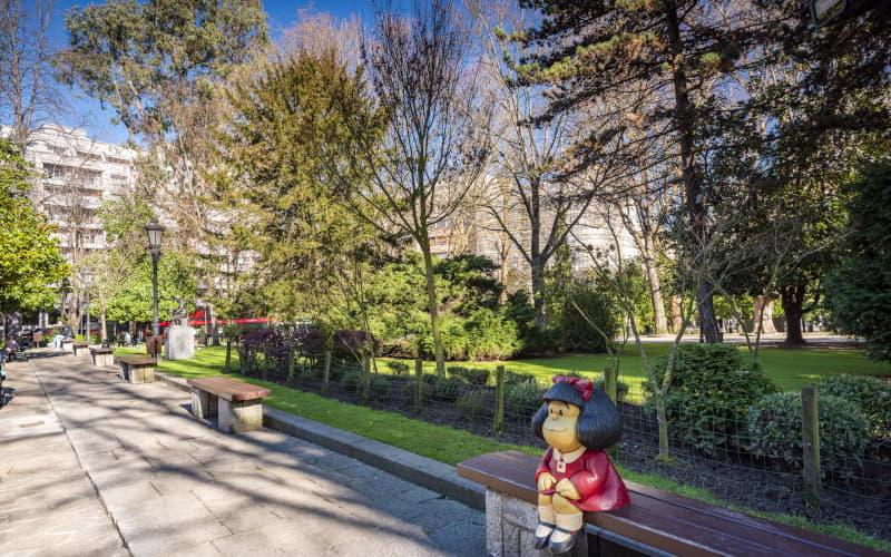 Estatua de Mafalda sentada en un banco del Parque de San Francisco en Oviedo, rodeada de árboles frondosos con edificios que asoman al fondo entre la vegetación.