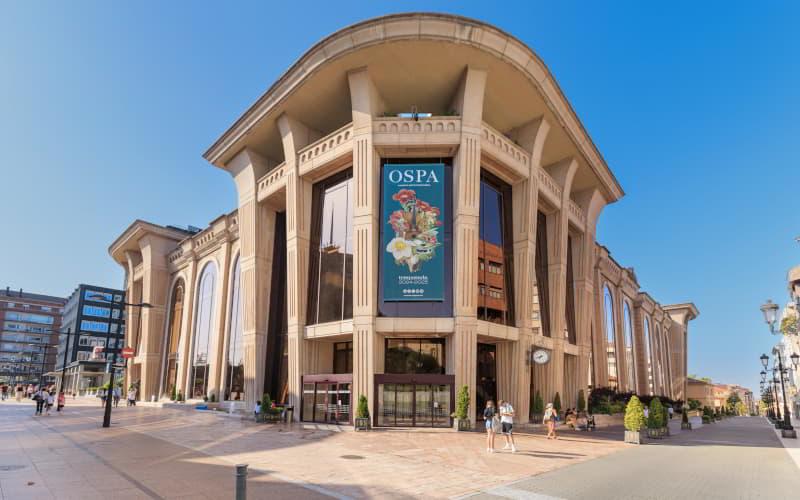 Edificio del Auditorio‑Palacio de Congresos Príncipe Felipe en Oviedo, visto desde una calle peatonal despejada bajo un cielo azul. La fachada combina elementos de aire neoclásico con gruesas columnas de hormigón arquitectónico y amplios ventanales que integran el agua como motivo decorativo