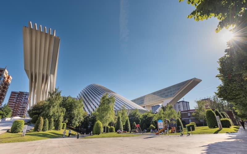 Edificio del Palacio de Exposiciones y Congresos de Oviedo, obra de Santiago Calatrava, emergiendo por encima de un parque infantil en un día soleado; su silueta presenta una estructura neofuturista de hormigón, acero y cristal, rematada por una gran cubierta de costillas blancas