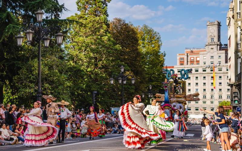 Desfile del Día de América en Asturias en la calle, con bailarines vestidos con trajes típicos coloridos de América Latina participando en la cabalgata; detrás se ven carrozas y público animando, rodeado de edificios y zonas verdes.