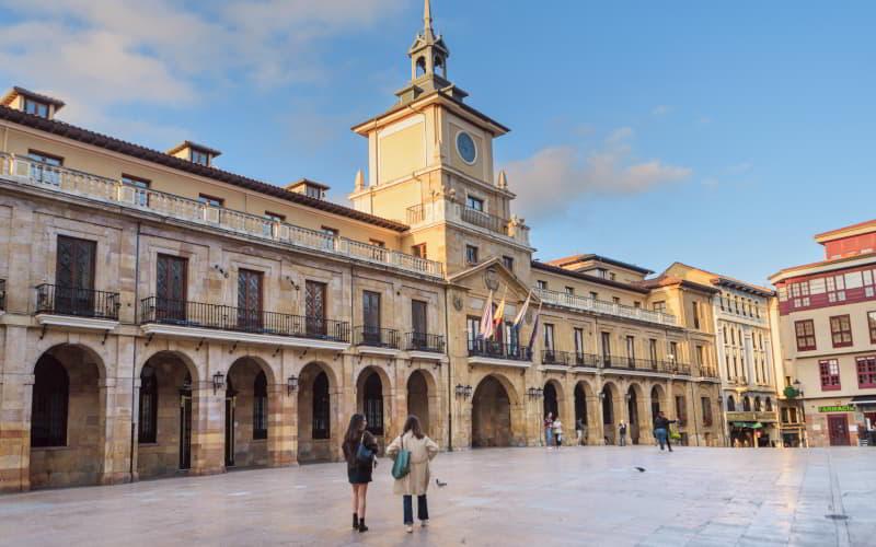 Plaza de la Constitución de Oviedo con el Ayuntamiento (Casa Consistorial) al fondo: edificio histórico de estilo barroco clásico con torre del reloj instalada en 1940, soportales en su planta baja y balcones con banderas; en el centro de la plaza, dos chicas conversan de pie bajo un cielo azul despejado.