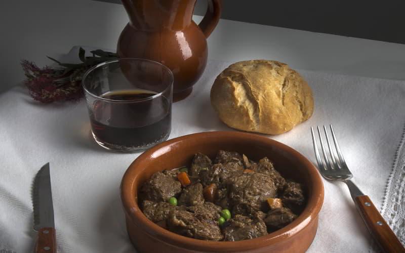Plato de barro con carne gobernada sobre una mesa con mantel blanco, acompañado de un bollo de pan redondo, un vaso de vino tinto y una jarra de barro; a los lados se disponen un tenedor y un cuchillo con mango de madera.