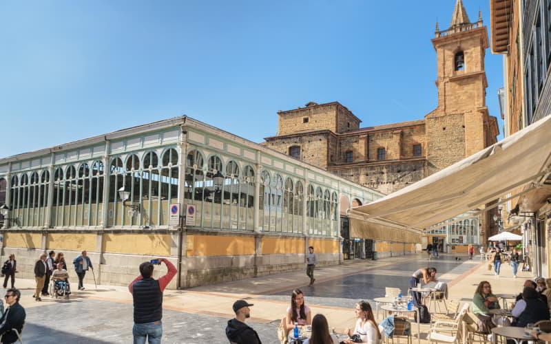 Vista del casco antiguo de Oviedo hacia la plaza del Fontán y sus calles peatonales de suelo liso; al fondo se alzan la iglesia de San Isidoro, mientras en primer plano la gente alterna en las terrazas y un turista fotografía la iglesia.