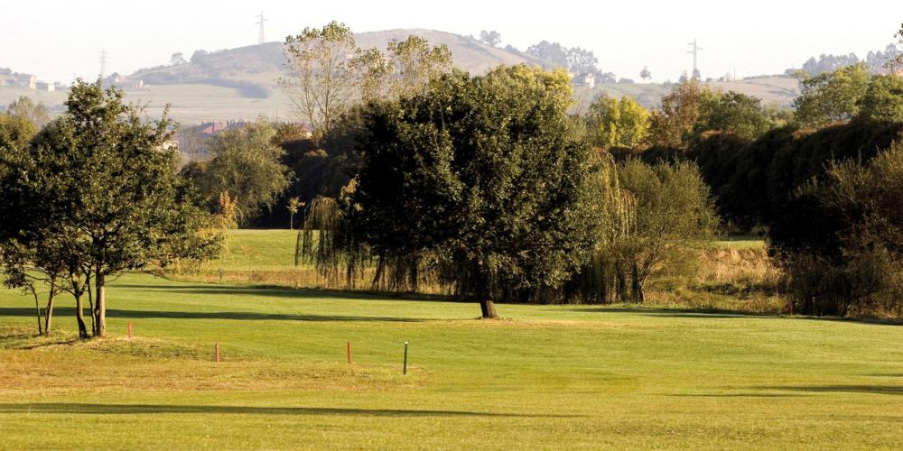 La Morgal golf course in Llanera, with lawns in the foreground with coloured tees embedded in the terrain; in the background, trees and a mountain landscape in shades of green and gold.