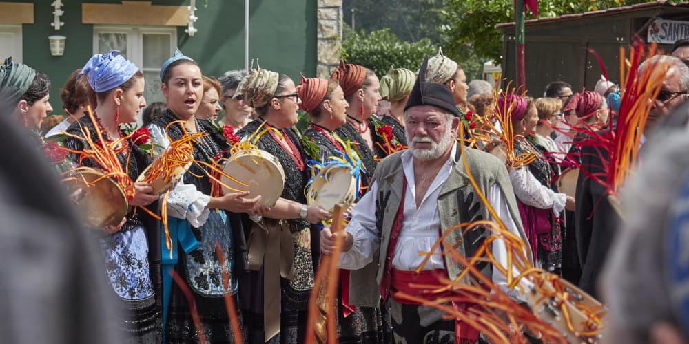 Bando Llanisco en las fiestas del Santo Cristo del Amparo, con un grupo de mujeres en fila cantando y tocando panderetas adornadas con lazos de colores de las banderas de España y de Asturias; entre la multitud avanza un hombre con atuendo regional, camisa blanca y cinturón rojo, apoyado en un objeto de madera, mientras al fondo se distingue una casa verde.