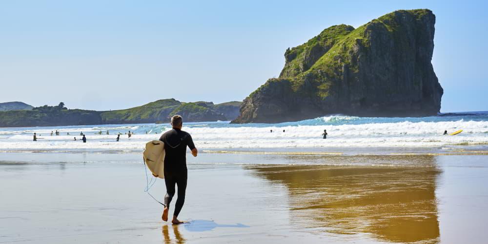 Surfista entrando al mar en la playa de San Martín, Llanes, con tabla en mano y traje de neopreno; al fondo se aprecian bañistas en el agua y un gran islote rocoso cubierto de vegetación.