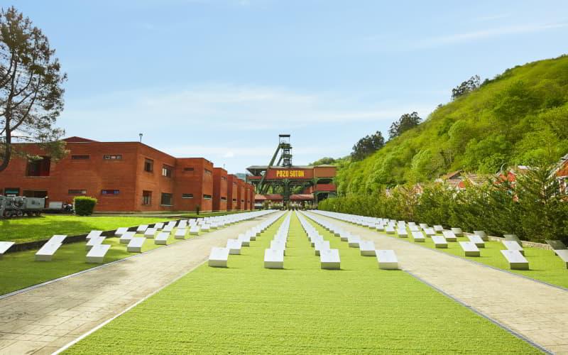 Memorial minero en el Pozo Sotón, San Martín del Rey Aurelio, con hileras de bloques blancos alineados en el espacio ajardinado que conduce a las instalaciones mineras de ladrillo rojo y a la emblemática torre de extracción al fondo.