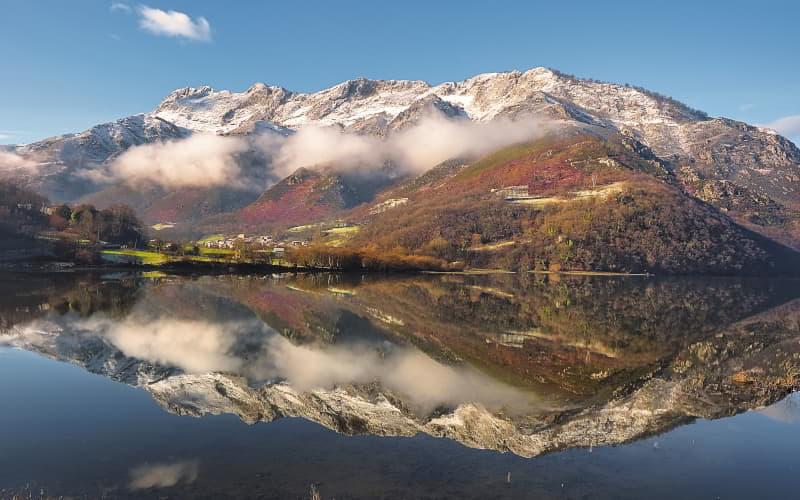 Embalse de Rioseco en Sobrescobio, con reflejo en el agua de un paisaje de montaña parcialmente nevada, un pueblo con casas rodeadas de árboles y vegetación en tonos otoñales de rojos y verdes intensos.