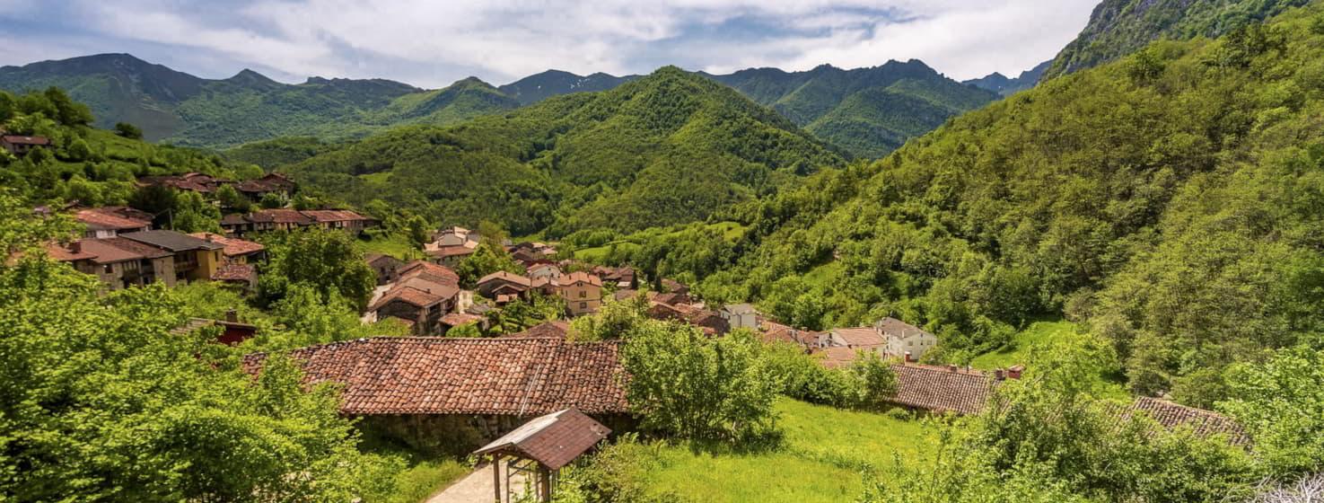 Vista panorámica de la aldea de Caleao, en el Parque Natural de Redes, con casas de piedra y tejados de teja rojiza rodeadas de montañas verdes y bosques, bajo un cielo parcialmente despejado.