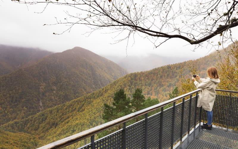 Chica con chubasquero en el mirador de Muniellos tomando una fotografía del paisaje, apoyada sobre una superficie tras una barandilla metálica perforada; al fondo se distingue un entorno nuboso y con niebla que deja entrever valles y montañas cubiertas de vegetación en tonos verdes y ocres.