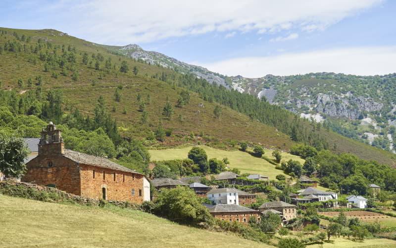 Vista del pueblo de Seroiro en Ibias, con una iglesia de piedra en primer plano y varias casas tradicionales rodeadas de prados y árboles, al pie de montañas cubiertas de vegetación y formaciones rocosas bajo un cielo parcialmente despejado.