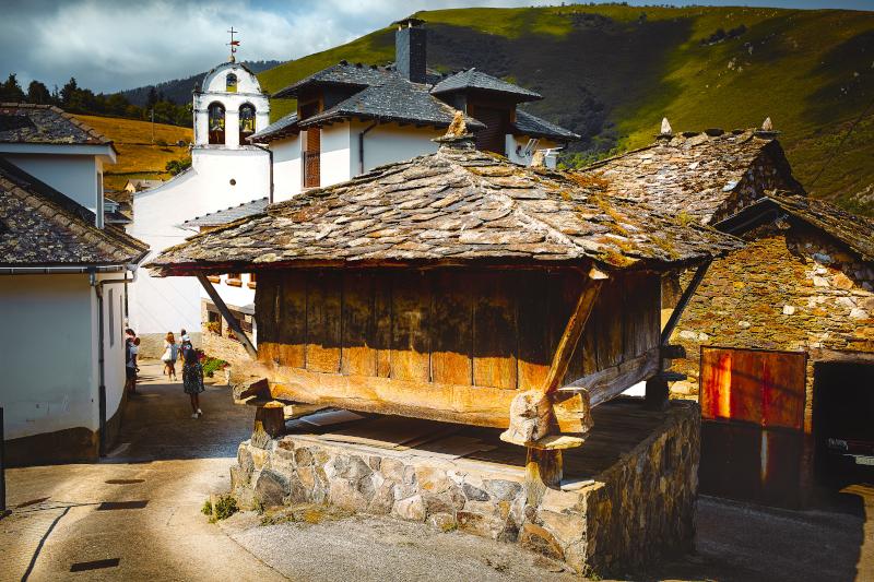 Vista de Besullo en Cangas del Narcea con un hórreo de piedra en primer plano junto a una calle pavimentada en pendiente; detrás del hórreo se distingue una casa blanca con tejado de pizarra gris y, más al fondo, una iglesia blanca con campanario de dos arcos. A la derecha, bajo la calle inclinada, se aprecia una construcción de piedra similar a un almacén con gran portón de madera y un coche en su interior. El conjunto está enmarcado por montañas cubiertas de verde bajo un cielo nublado.