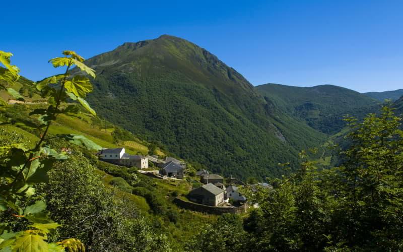 Vista de Brañas de Arriba desde lo alto, con un conjunto de casas en el centro rodeadas de prados y montañas, y al fondo el Cueto de Arbas bajo un cielo azul completamente despejado.