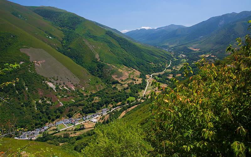 Vista panorámica de Fonduveigas en Degaña, con el pueblo en el fondo del valle rodeado de montañas verdes y laderas con zonas de bosque y claros, atravesado por una carretera que serpentea entre la vegetación bajo un cielo despejado.