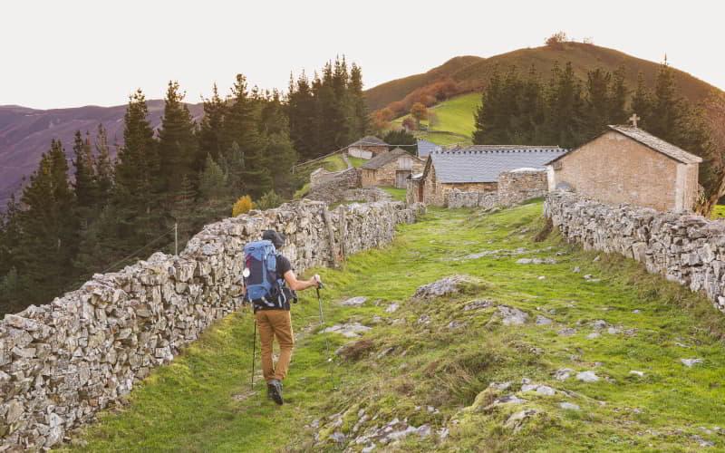 Pilger mit Rucksack und Wanderstöcken auf einem gepflasterten Weg des Primitiven Weges in Montefurado, umgeben von Steinmauern, traditionellen Häusern und waldbedeckten Bergen.
