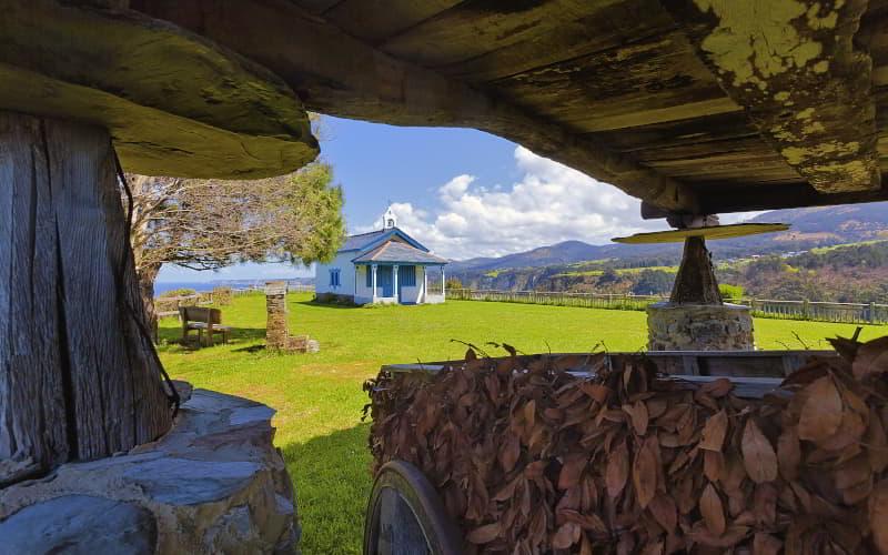 Blick auf die Einsiedelei La Regalina in Cadavéu, die auf einer grünen Wiese mit Bäumen und Steinbänken liegt, in einer offenen Umgebung mit Bergen im Hintergrund.