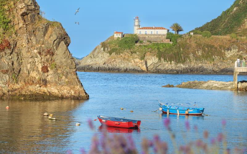 Blick auf den Leuchtturm von Cudillero, der auf einem Felsvorsprung steht, mit kleinen Fischerbooten, die in der Bucht schwimmen.