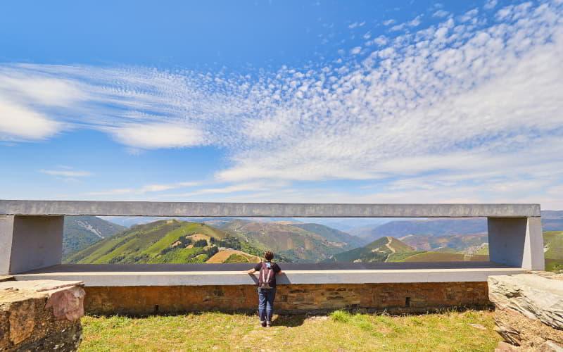 Eine Person, die die Landschaft vom Aussichtspunkt Montefurado aus betrachtet, mit einem weiten Blick auf vegetationsbedeckte Berge unter einem blauen Himmel mit vereinzelten Wolken.