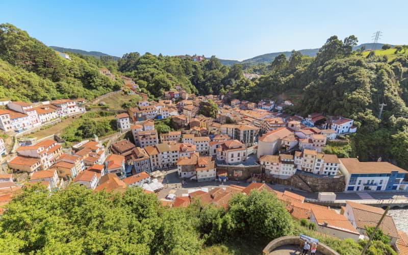 Panoramablick auf Cudillero mit seinen terrassenförmig in den Hang gebauten Häusern mit rötlichen Dächern, umgeben von bewaldeten Hügeln, von einem erhöhten Standpunkt aus gesehen.