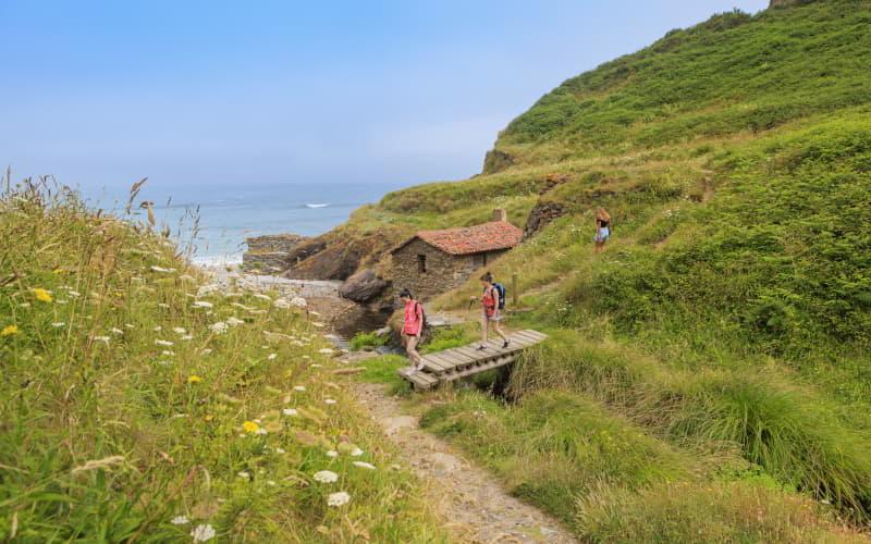 Küstenweg zum Strand von La Vallina, der über eine kleine Holzbrücke und eine alte Steinmühle mit rotem Dach direkt am Meer führt.