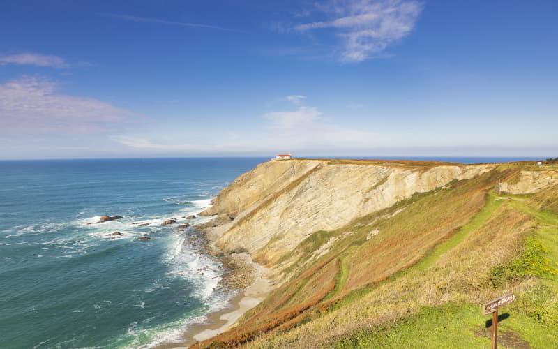 Die Klippen von Kap Vidio von einem Weg an der Küste aus gesehen, mit dem Kantabrischen Meer, das sich an den Felsen bricht, und einem Gebäude auf der Spitze des Vorgebirges.