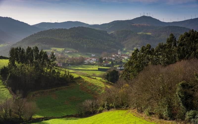 Panoramablick auf San Martín de Luiña von Mumayor aus, mit grünen Wiesen, Baumhainen und dem Weiler im Tal, umgeben von Bergen mit Windrädern auf den Gipfeln.