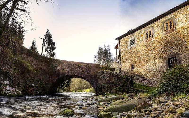 Puente de piedra de arco único sobre un río con corriente, rodeado de vegetación de ribera. A la derecha se alza una edificación tradicional de mampostería con ventanas pequeñas, bajo un cielo despejado en la parte superior.