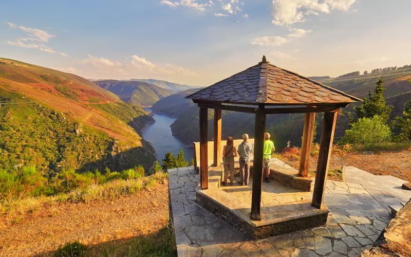 Mirador con tejadillo de pizarra y pilares de madera, desde el que varias personas contemplan el valle del río Navia. El paisaje muestra laderas montañosas con vegetación y el embalse serpenteando entre ellas bajo un cielo parcialmente despejado.