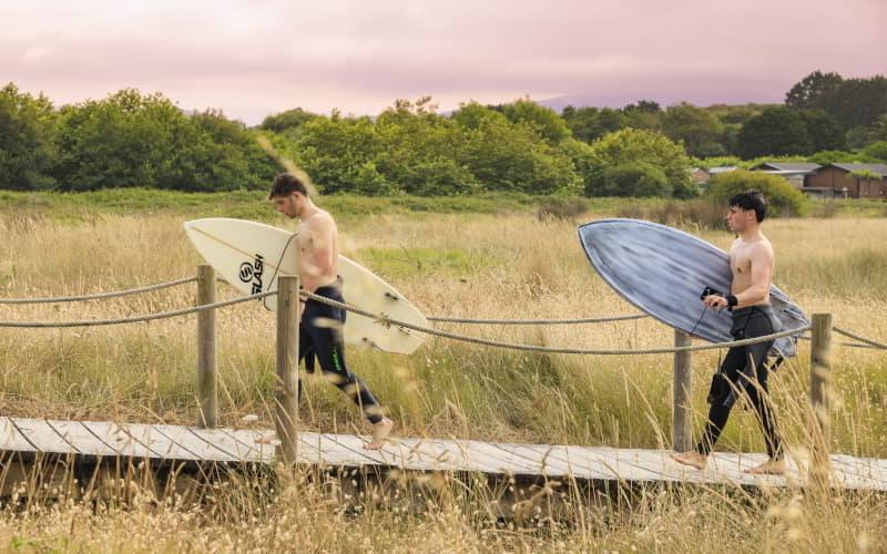 Dos surfistas caminan descalzos por una pasarela de madera entre dunas y vegetación, llevando sus tablas bajo el brazo en dirección a la playa. El entorno está rodeado de prados y árboles, con cielo cubierto al atardecer.