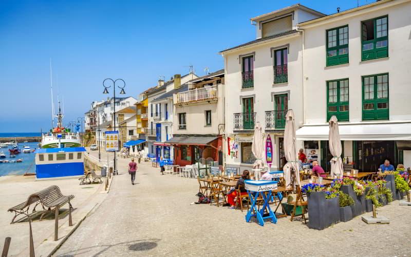 Vista del puerto de Tapia de Casariego con varias embarcaciones atracadas y una calle peatonal junto al muelle. A la derecha se alinean edificios de fachadas coloridas con terrazas de bares y restaurantes donde se sientan varias personas bajo sombrillas en un día soleado.