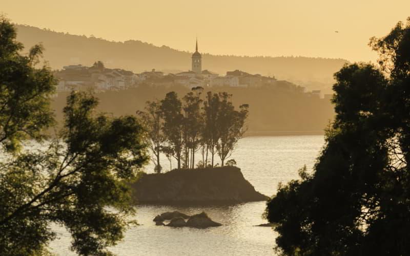 Pôr do sol no estuário do Eo, com uma pequena ilha arborizada em primeiro plano e a silhueta da cidade de Castropol e da sua igreja ao fundo, envolta em luz dourada.
