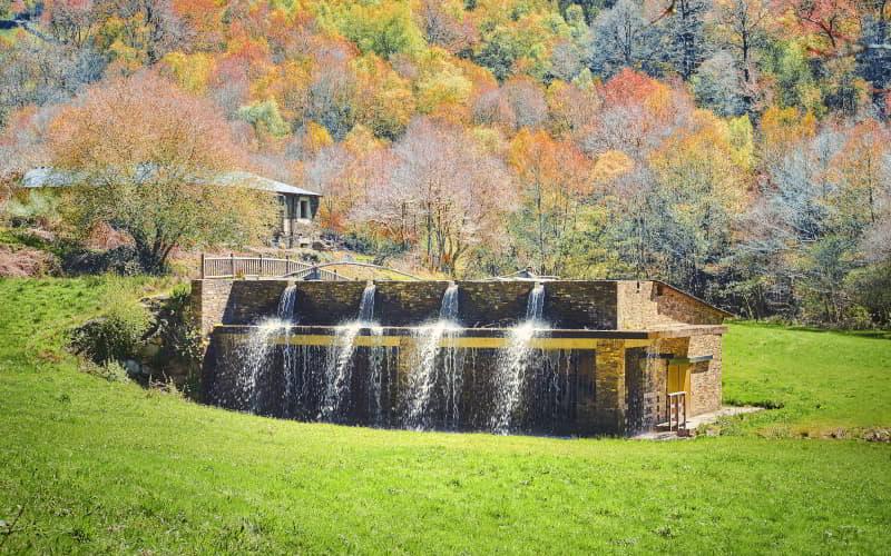 La Ferrería de Santa Eufemia em Villanueva de Oscos, um edifício de pedra com água a cair em cascata pela fachada, rodeado de prados e bosques com cores outonais.