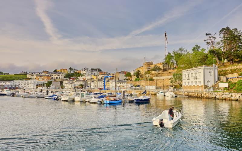 Barcos atracados no porto de Figueras, em Castropol, com um pequeno barco a navegar em primeiro plano e o centro da cidade na encosta ao fundo, sob um céu parcialmente nublado.