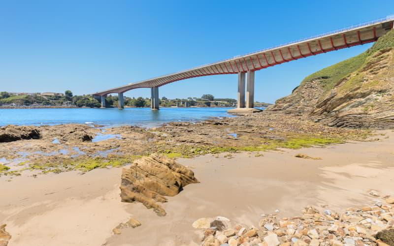 Vista da ponte sobre o rio Eo a partir da praia de San Román em Castropol, com areia, rochas e zonas de maré baixa em primeiro plano, sob um céu limpo.