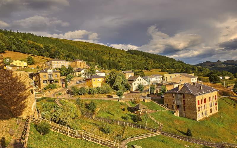 Vista do centro de Santa Eulália de Oscos, com casas de pedra e telhados de ardósia em encostas verdes, rodeadas por florestas e montanhas, à luz quente da tarde.