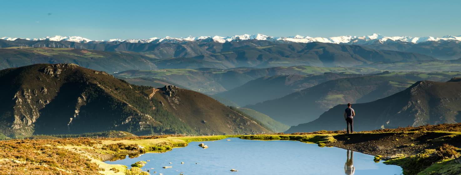 Vista panorâmica de Villanueva de Oscos mostrando uma paisagem montanhosa de La Bobia com um céu limpo. Em primeiro plano, uma piscina de água reflecte o ambiente, enquanto um homem contempla a vista. Ao fundo, uma cadeia de montanhas cobertas de neve estende-se sobre um horizonte de vales e colinas, criando um belo contraste entre o verde e o branco.