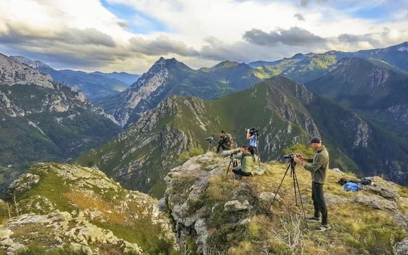Grupo de personas con cámaras y trípodes observa la fauna desde un mirador natural en una cima rocosa de Proaza. El paisaje muestra un amplio valle rodeado de montañas y bosques bajo un cielo parcialmente nublado con luz de tarde.