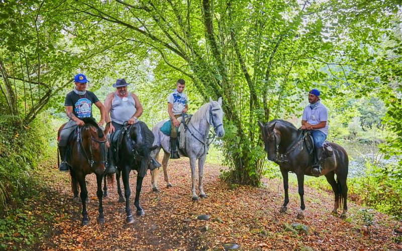 Cuatro jinetes montan a caballo por un sendero arbolado cubierto de hojas secas en el Camino Primitivo, en Las Regueras. El entorno muestra vegetación densa y un ambiente húmedo, con el río visible al fondo entre los árboles.