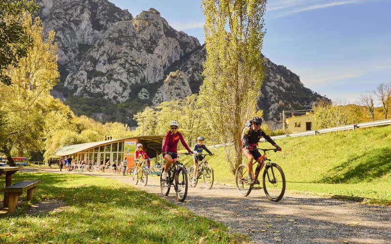 Grupo de ciclistas recorre un camino junto al embalse de Valdemurio, rodeado de praderas y árboles en un entorno montañoso. Al fondo se aprecian paredes rocosas y un edificio con zona de descanso bajo cielo despejado y luz diurna.