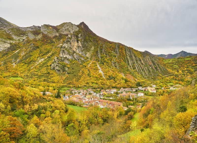 Vista panorámica de La Pola de Somiedo rodeada de montañas con laderas cubiertas de bosques otoñales en tonos amarillos y verdes. El núcleo urbano se asienta en el fondo del valle bajo un cielo nublado.
