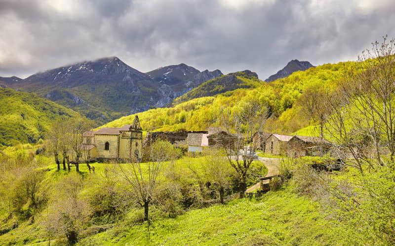 Vista del pequeño núcleo rural de Lindes, con una iglesia de piedra y varias casas tradicionales rodeadas de praderas y árboles. El paisaje se enmarca entre montañas con cumbres parcialmente nevadas y un cielo cubierto.