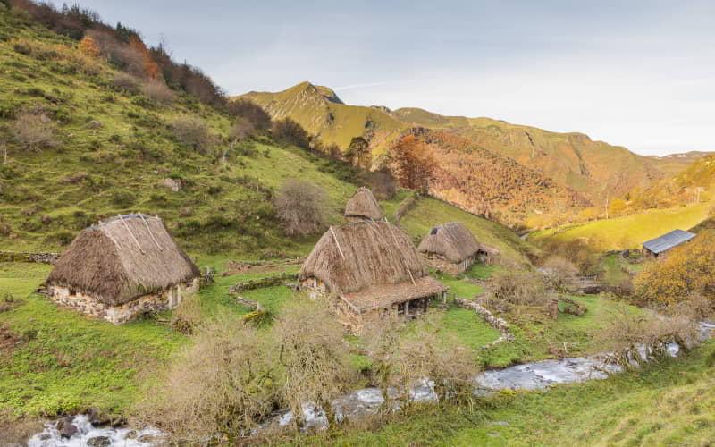 Conjunto de construcciones tradicionales con techos de escoba en la braña de La Pornacal, dentro del Parque Natural de Somiedo. Las cabañas se ubican junto a un arroyo entre praderas y laderas de montaña cubiertas de vegetación, bajo luz suave de tarde.