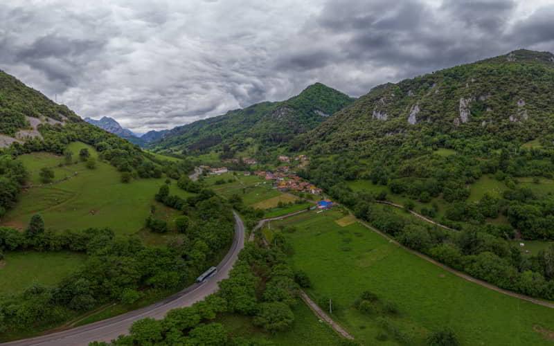 Vista aérea del valle del río Trubia con el pueblo de Villanueva en el centro, rodeado de montañas cubiertas de vegetación. El paisaje muestra praderas verdes, una carretera serpenteante y un cielo nublado que cubre parcialmente las cumbres del entorno.