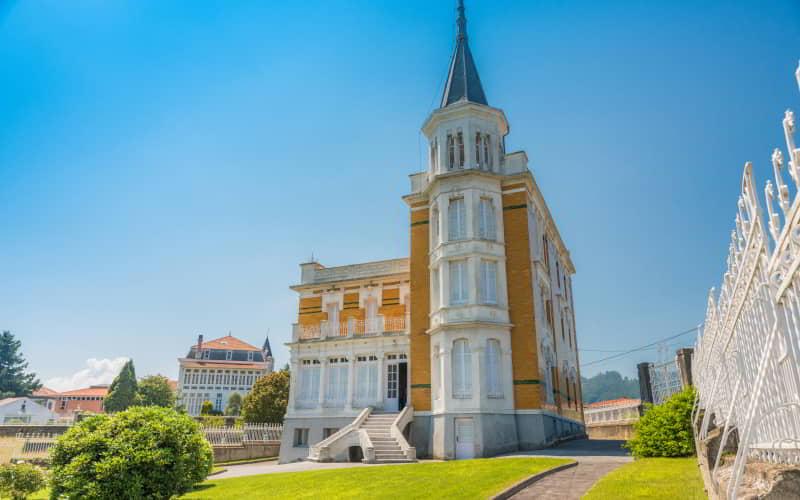 Edificio histórico de estilo ecléctico con fachada de ladrillo y torre esquinera rematada en aguja metálica. Se encuentra rodeado por un jardín cuidado y una verja blanca, bajo un cielo despejado y luminoso.