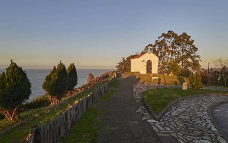 Pequeña ermita blanca situada en un acantilado junto al mar, rodeada de árboles y vegetación baja. Un camino pavimentado conduce hasta el mirador, iluminado por la luz cálida del atardecer.