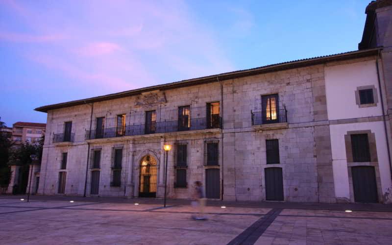 Fachada principal del Palacio de los Moutas, edificio histórico de piedra con balcones de hierro forjado y un portal central ornamentado. La escena muestra la plaza al anochecer, con iluminación cálida y un cielo azul rosado al fondo.