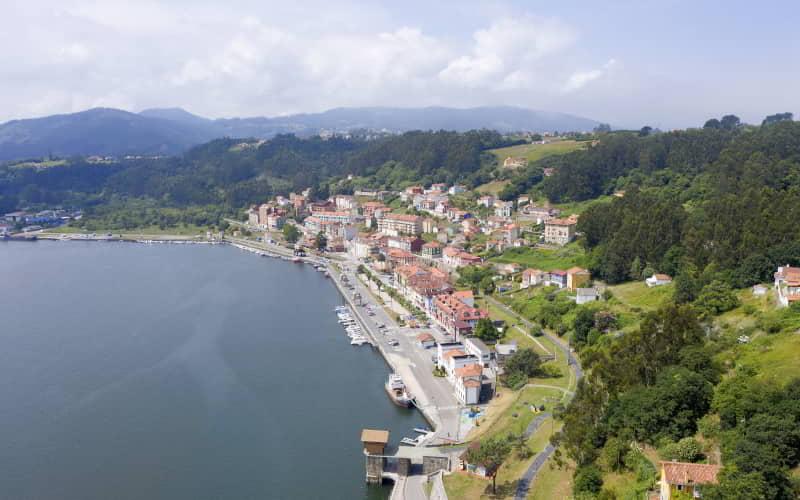 Vista aérea del puerto de San Esteban de Pravia, con el paseo marítimo y las antiguas estructuras de carga de carbón junto al río Nalón. Las casas se disponen en terrazas sobre la ladera verde, con colinas boscosas y montañas suaves al fondo bajo un cielo parcialmente nublado.