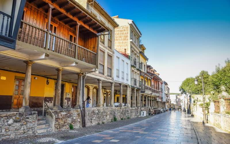 Vista da Calle Galiana em Avilés, com arcadas de pedra e fachadas tradicionais de cores quentes e varandas de madeira. O pavimento molhado reflecte a luz do dia e a rua parece quase vazia sob um céu limpo.