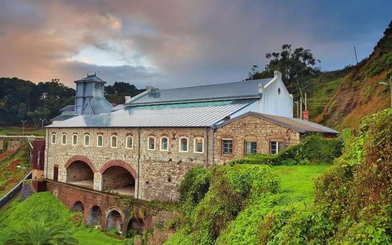 Edifício de pedra com estrutura metálica que alberga o Museu da Mina de Arnao, situado numa encosta coberta de vegetação. O edifício assenta em arcos de tijolo junto a uma encosta verde, com céu parcialmente nublado e luz quente ao entardecer.