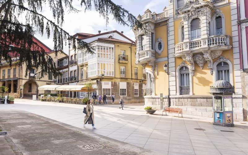 Fachada do Palácio de Balsera em Avilés, um edifício histórico de estilo eclético com pormenores ornamentais e varandas de pedra. Em primeiro plano, uma praça pedonal com pessoas a passear e esplanadas de cafés com uma suave luz do dia.